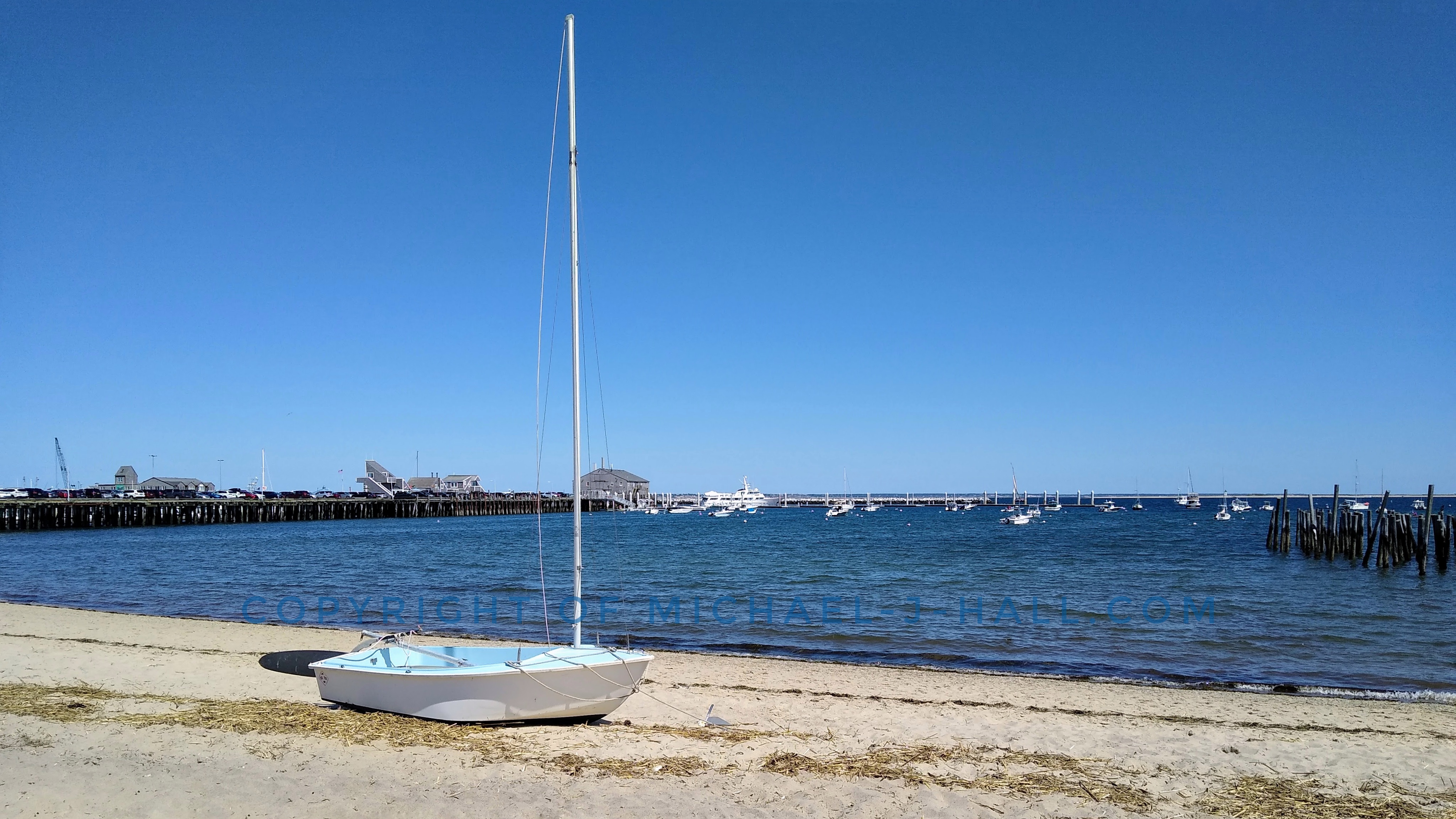 On this clear, cool day a masted skiff is anchored loosely in the sandy beach ready to bravely challenge the Provincetown bay whenever its owner returns for their next bold adventure with the wind and water together.